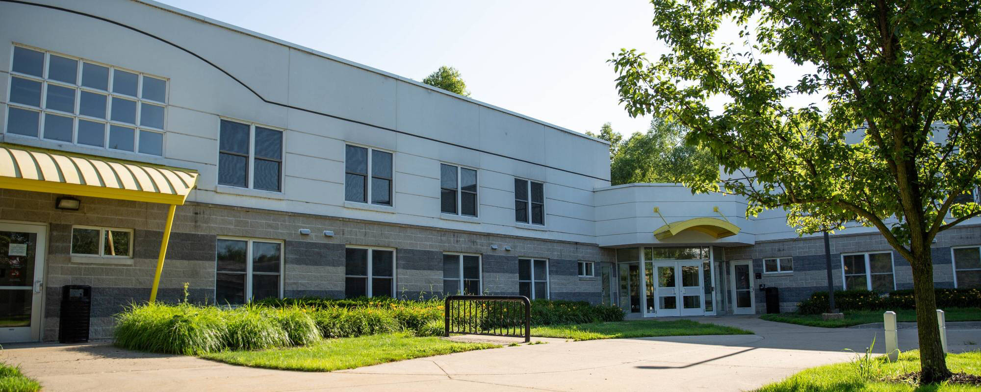 A modern two-story school building, Calder Residence Center, with large windows, a yellow entrance awning, and landscaped greenery.